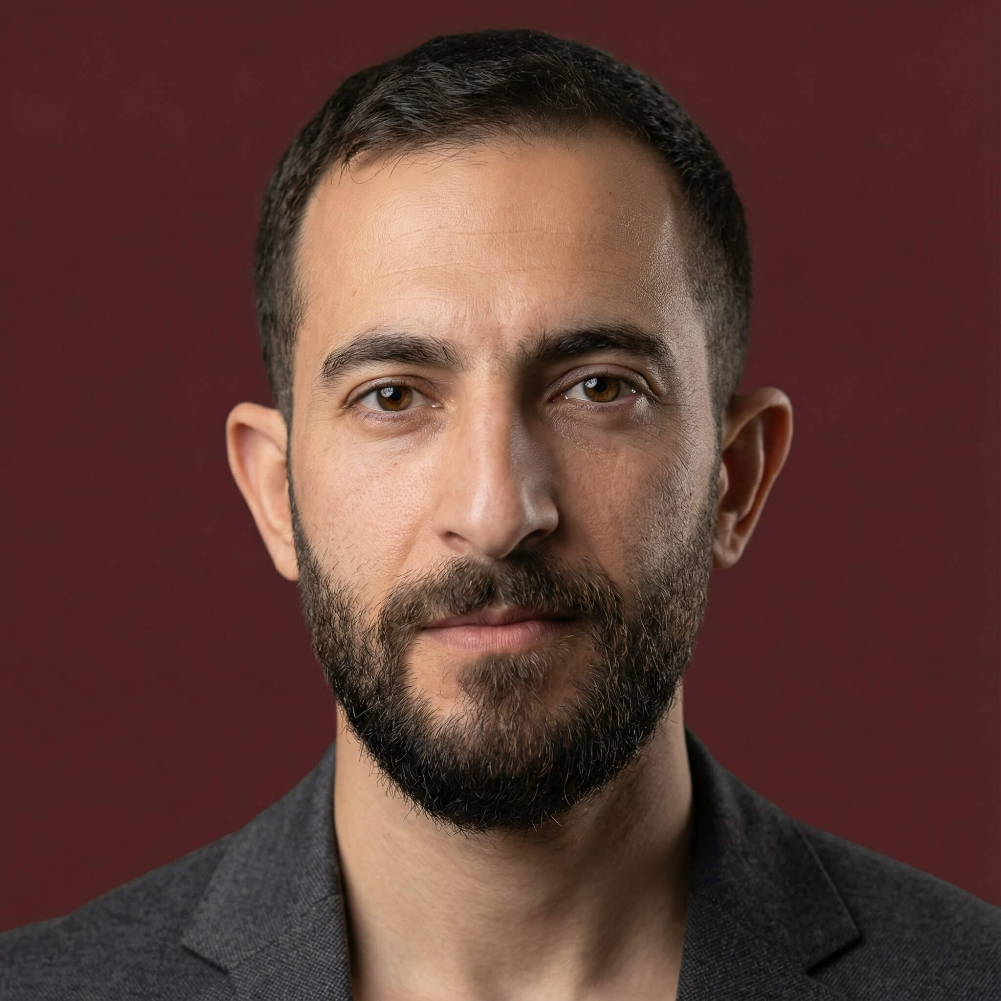 Studio headshot of a person with a short beard on a dark red background